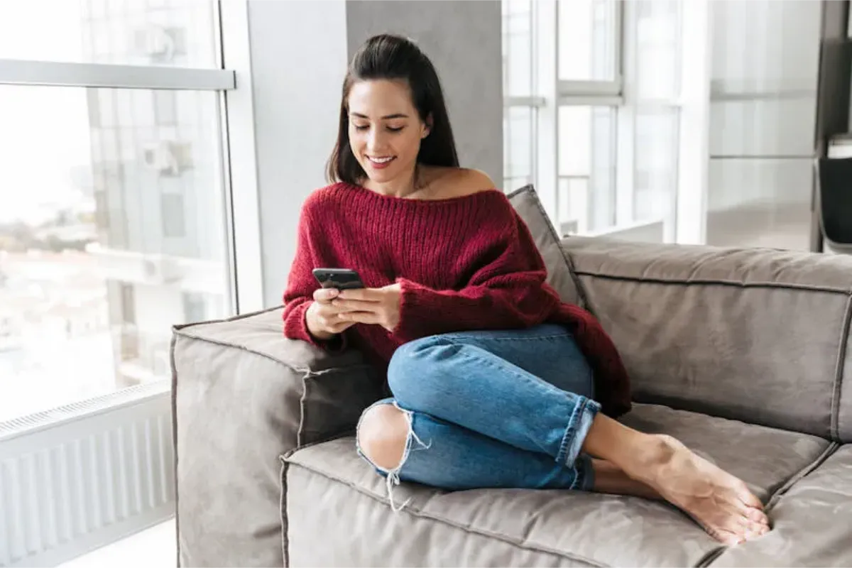 Brunette woman smiling and sitting on a sofa texting and chatting on her phone