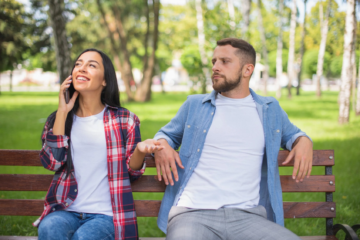 Couple sitting on a park bench, girl is happy and chatting on the phone while guy looks unhappy